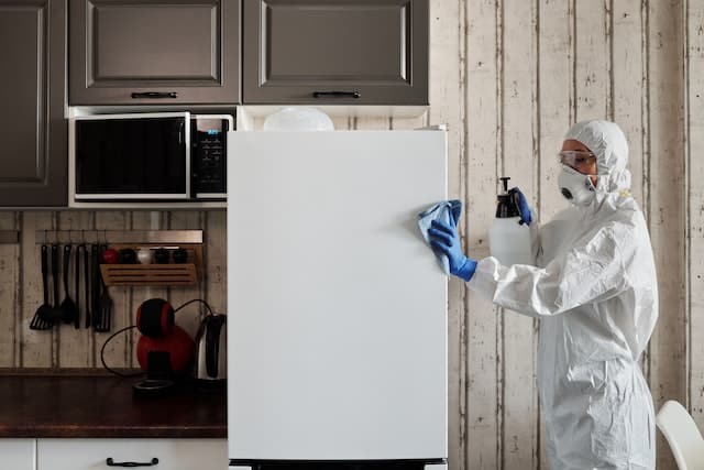 a person dressed in protective cleaning clothing and a mask with a rag and bottle of cleaning liquid cleaning a white fridge in a kitchen