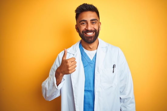 Young indian doctor man standing over isolated yellow background doing happy thumbs up gesture with hand. Approving expression looking at the camera with showing success.
