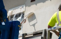 Builder,Painting,A,Building,Exterior,In,Bright,Sunlight,While,On Builder painting a building exterior in bright sunlight while on a lift