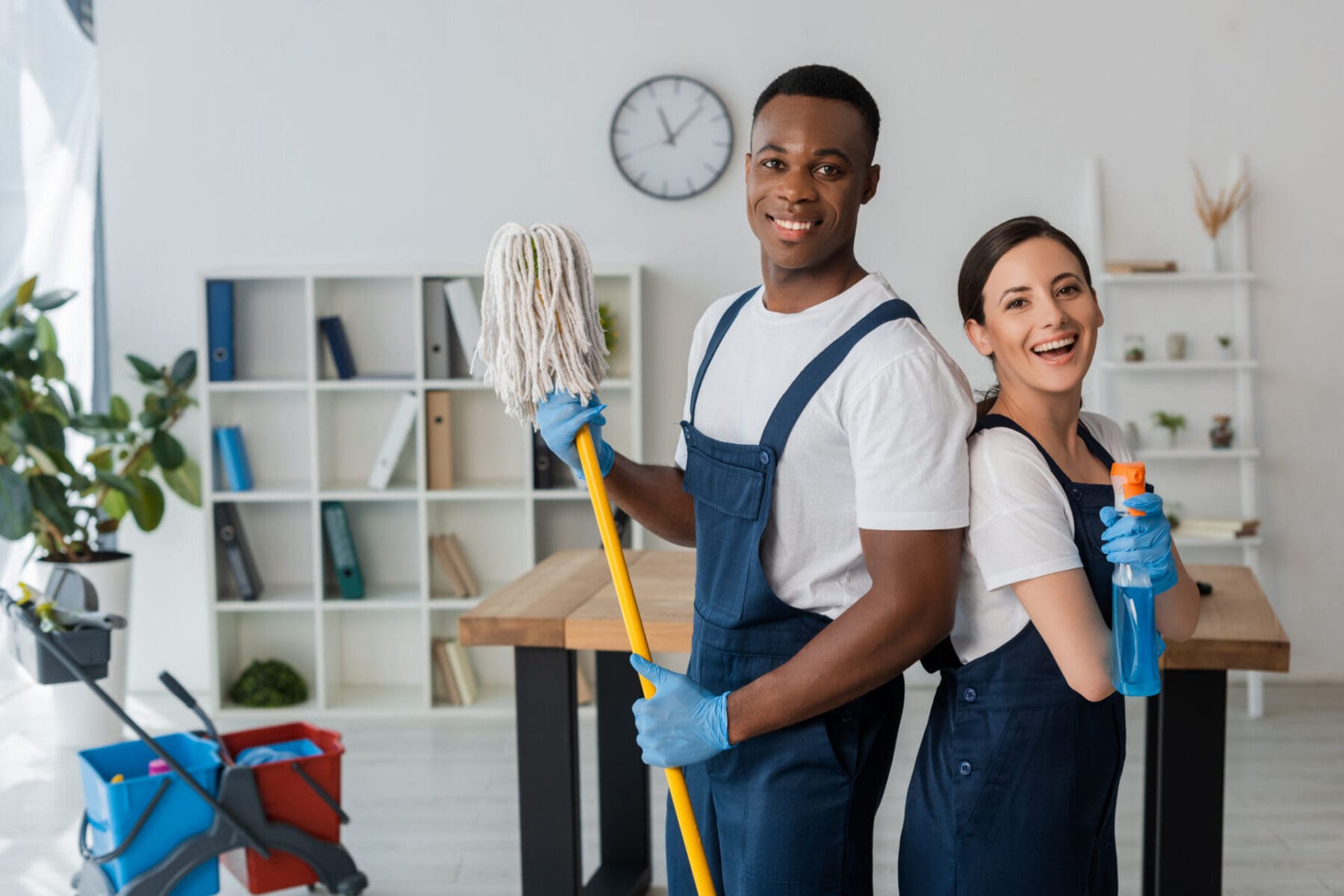 Positive multiethnic cleaners holding mop and detergent while smiling at camera in office janitorial crew