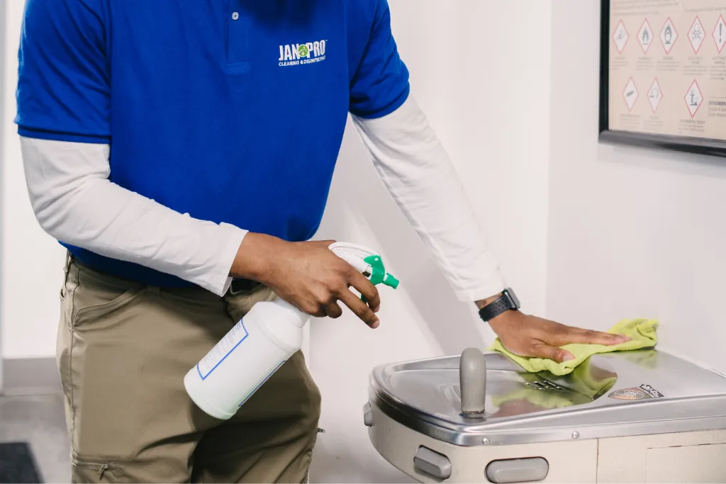 A cleaning professional sprays disinfectant and wipes a metal drinking fountain with a cloth.
